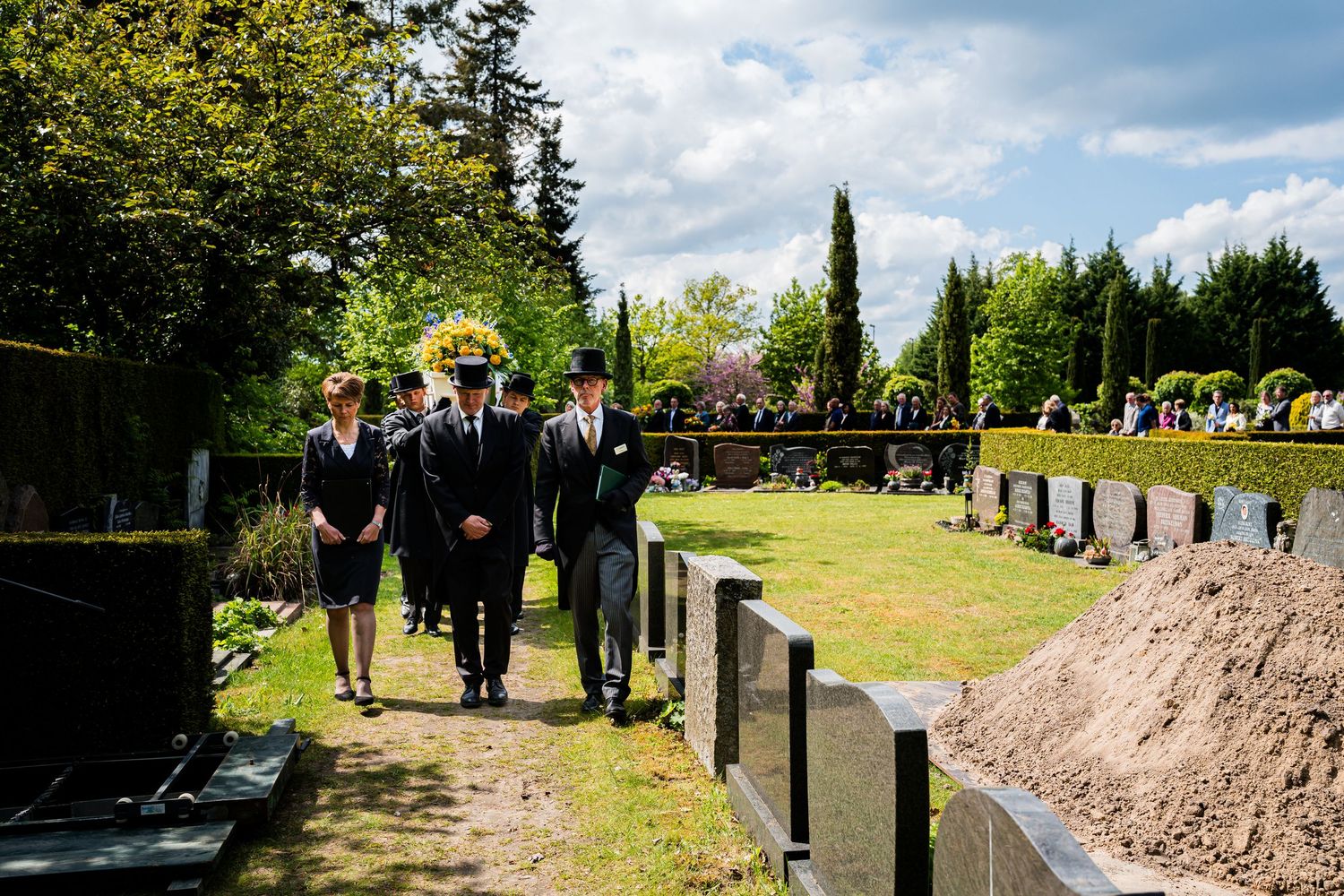 Funeral procession walking through a cemetery on a sunny day, with mourners in formal black attire carrying a floral wreath.