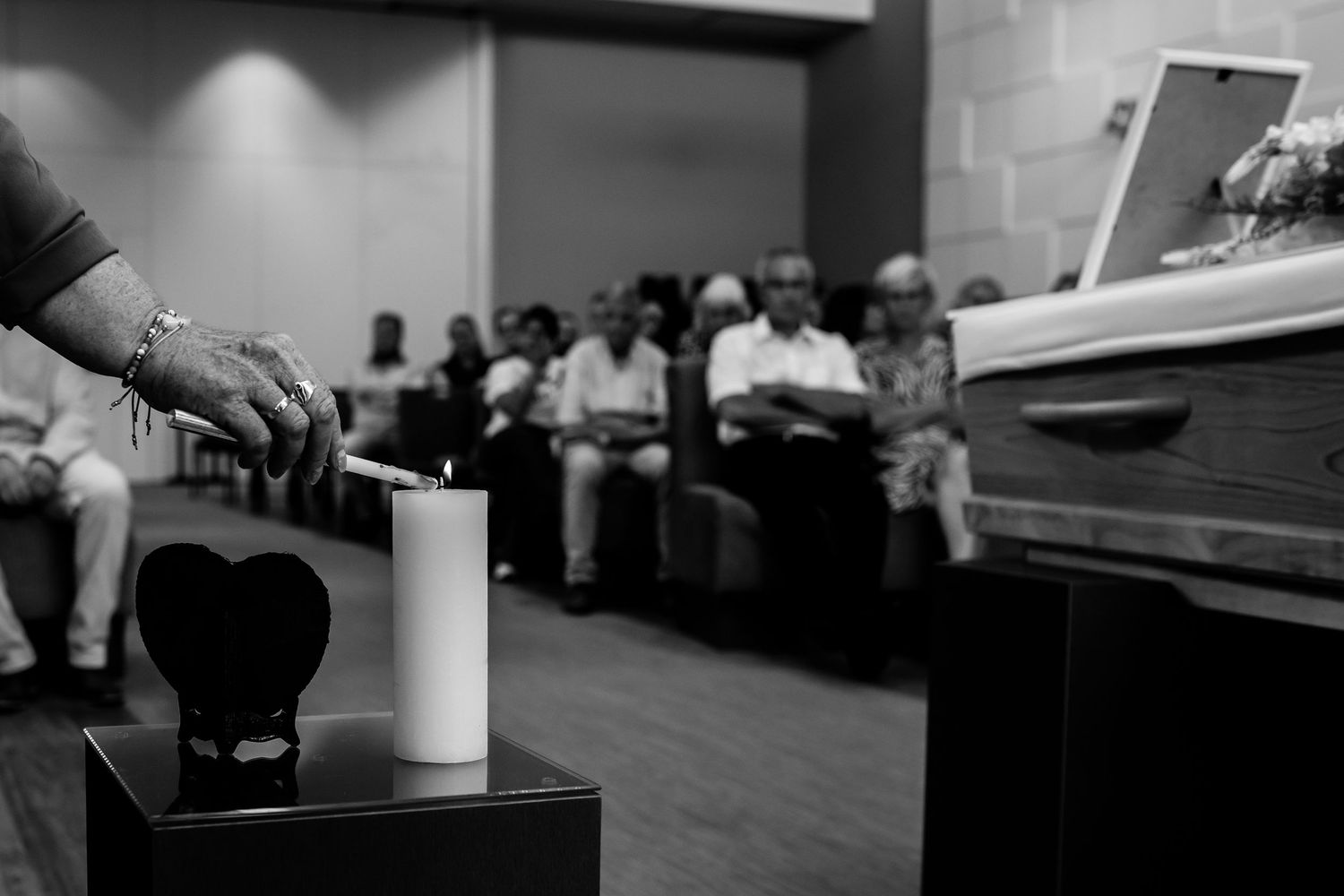 Black and white funeral service with mourners seated, a lit candle, heart urn, and casket in a somber chapel.