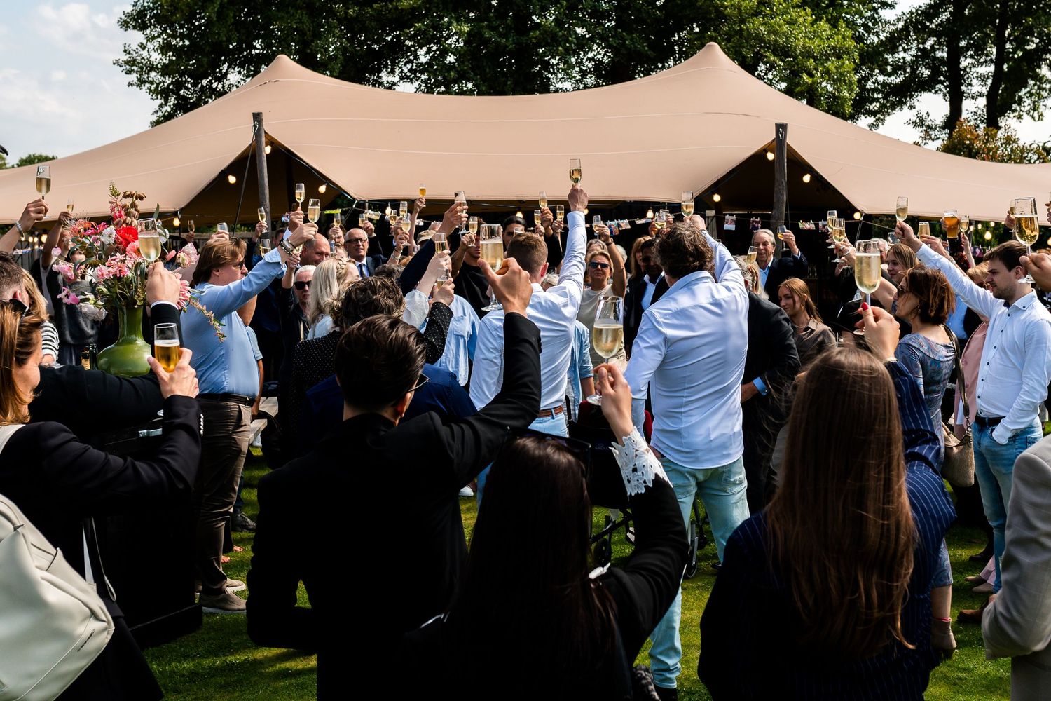 Guests raising glasses in a toast outdoors at a wedding reception with a large marquee tent in the background.
