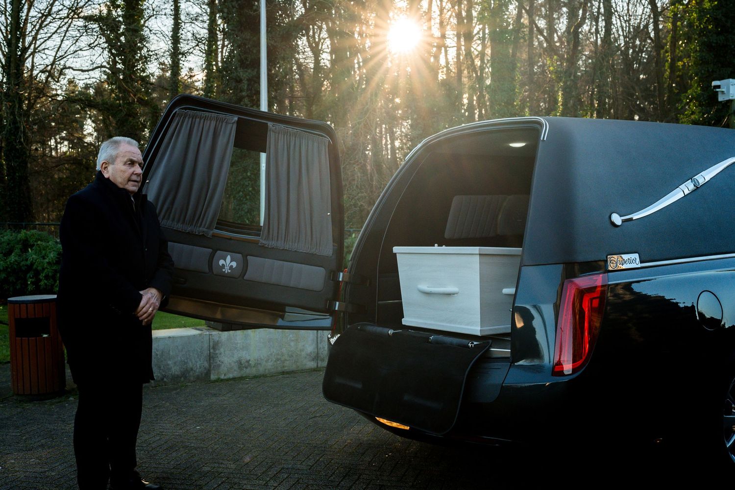 Elderly man standing beside a hearse with white casket at sunset in a peaceful outdoor setting.