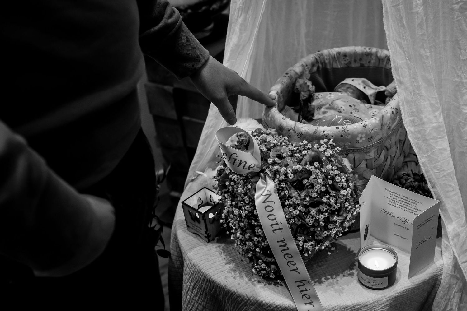 Black and white photo of a baby in a flower-adorned bassinet with candle and memorial items nearby.