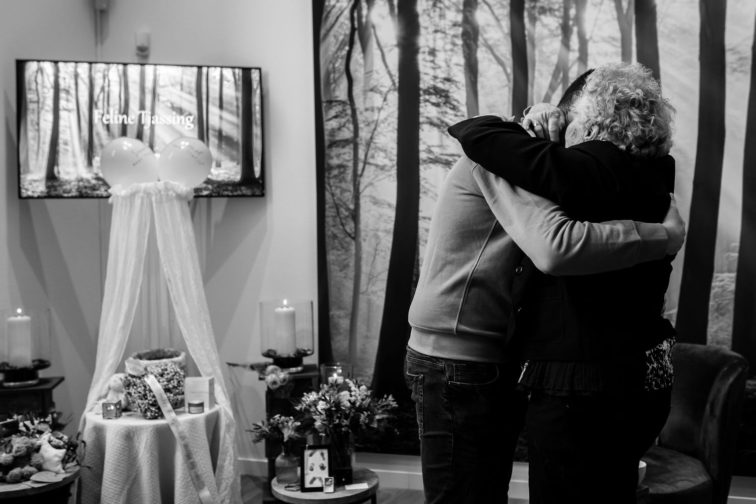 Two people share a comforting embrace at a memorial service with floral arrangements and decorations in a black and white photo.
