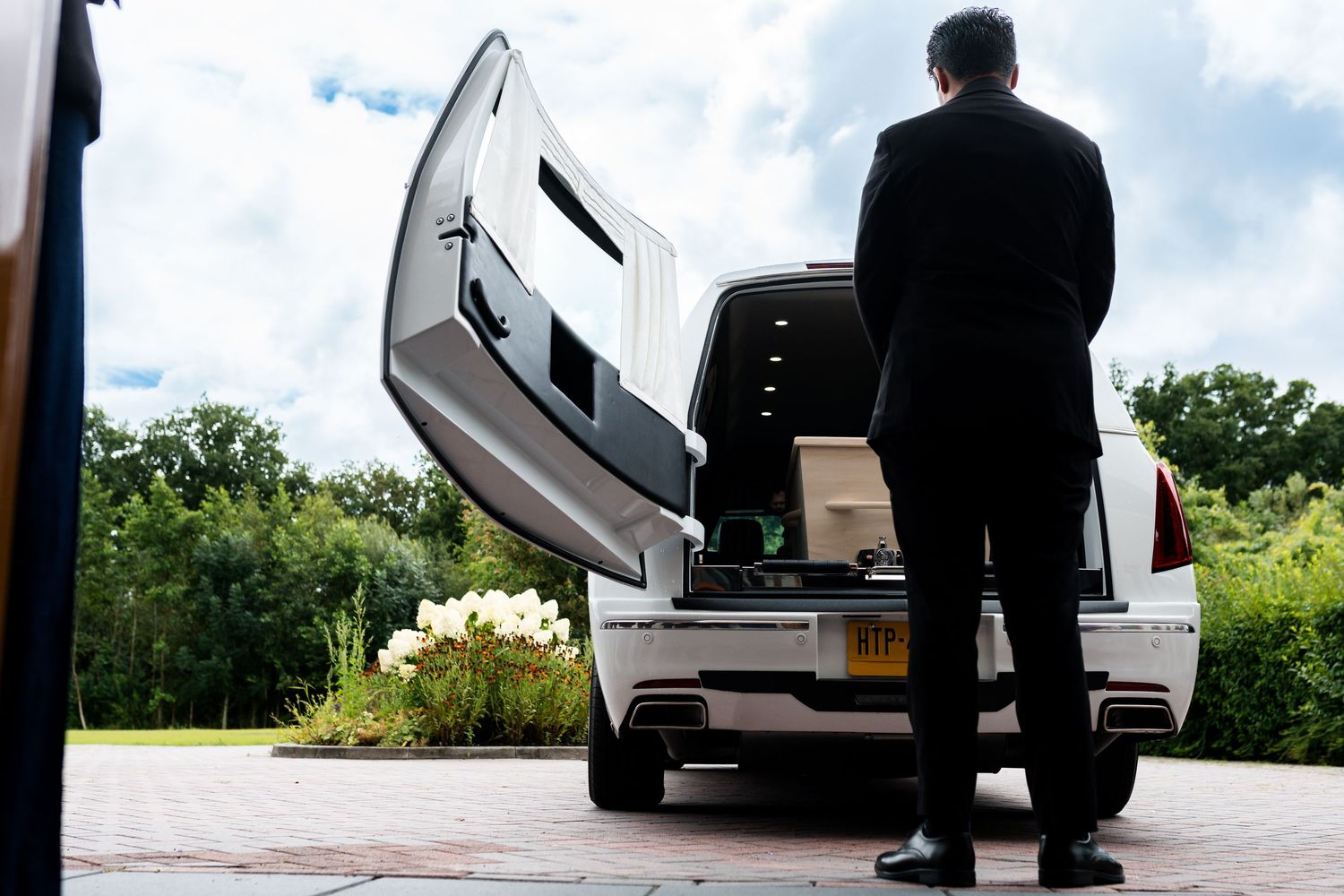 Funeral director standing beside an open hearse with a casket, surrounded by lush greenery on a sunny day.