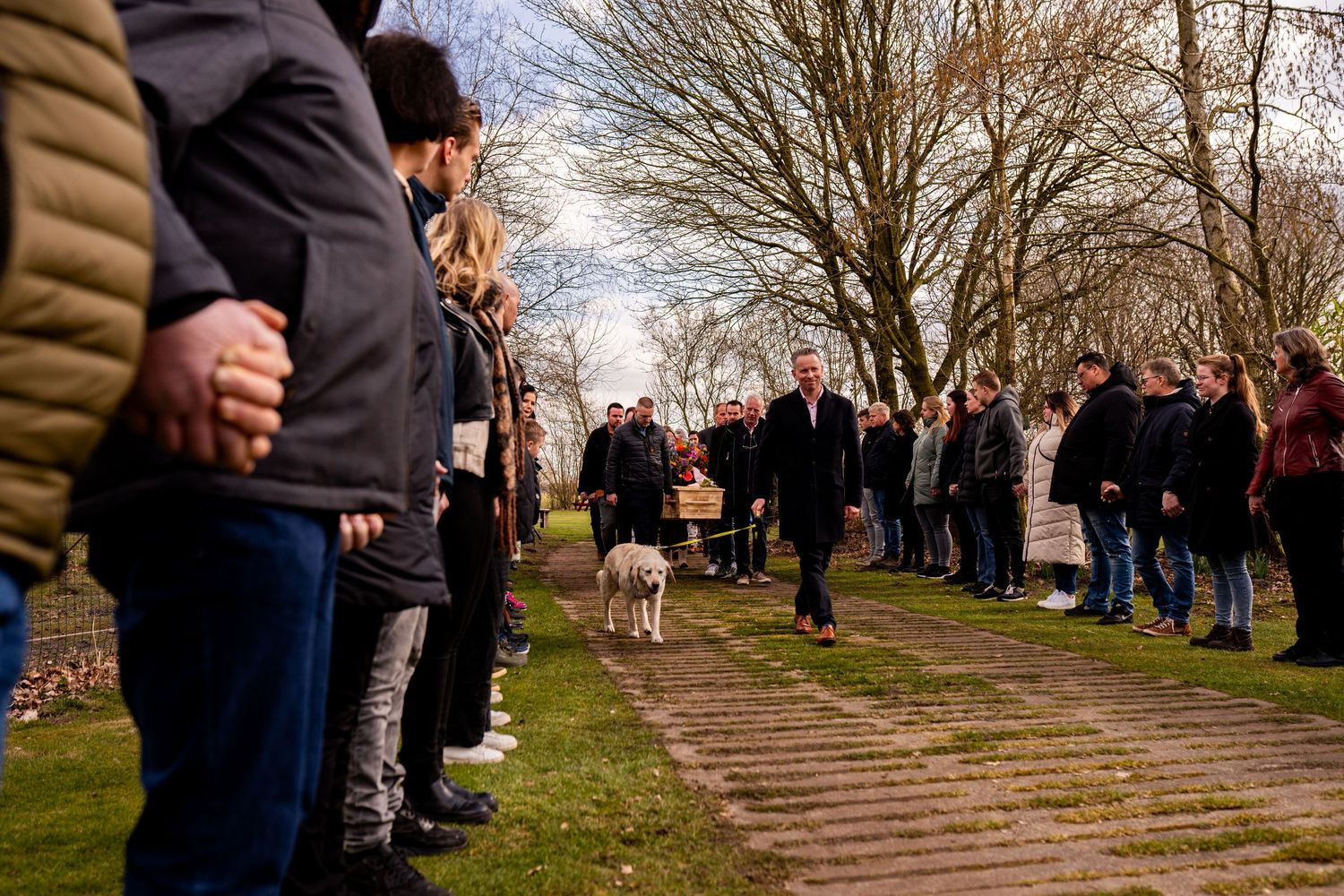 People lined up outdoors along a path as a man walks a white dog during a somber gathering in a park.