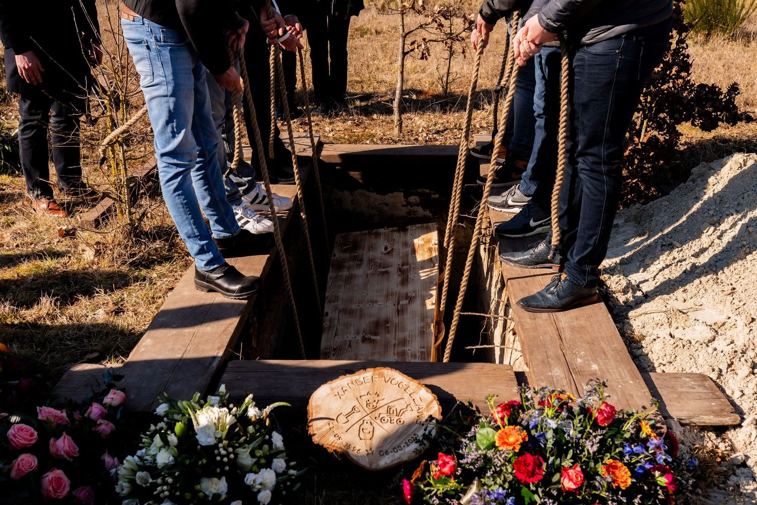 People standing around an open grave with flowers at a funeral burial ceremony outdoors.