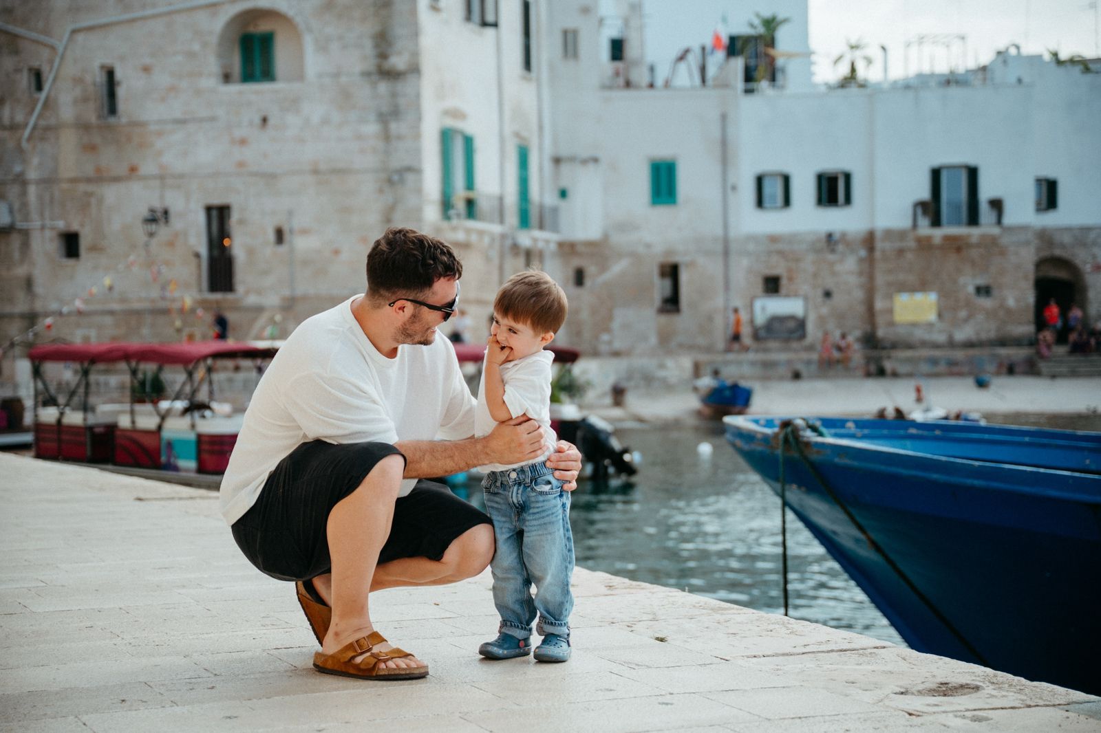 Father and toddler son bonding on sunny harbor dock beside blue boat in historic town.