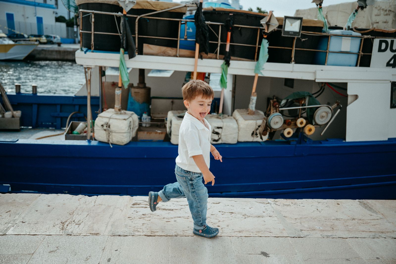 A young toddler boy running along a harbor dock with a blue fishing boat in the background.