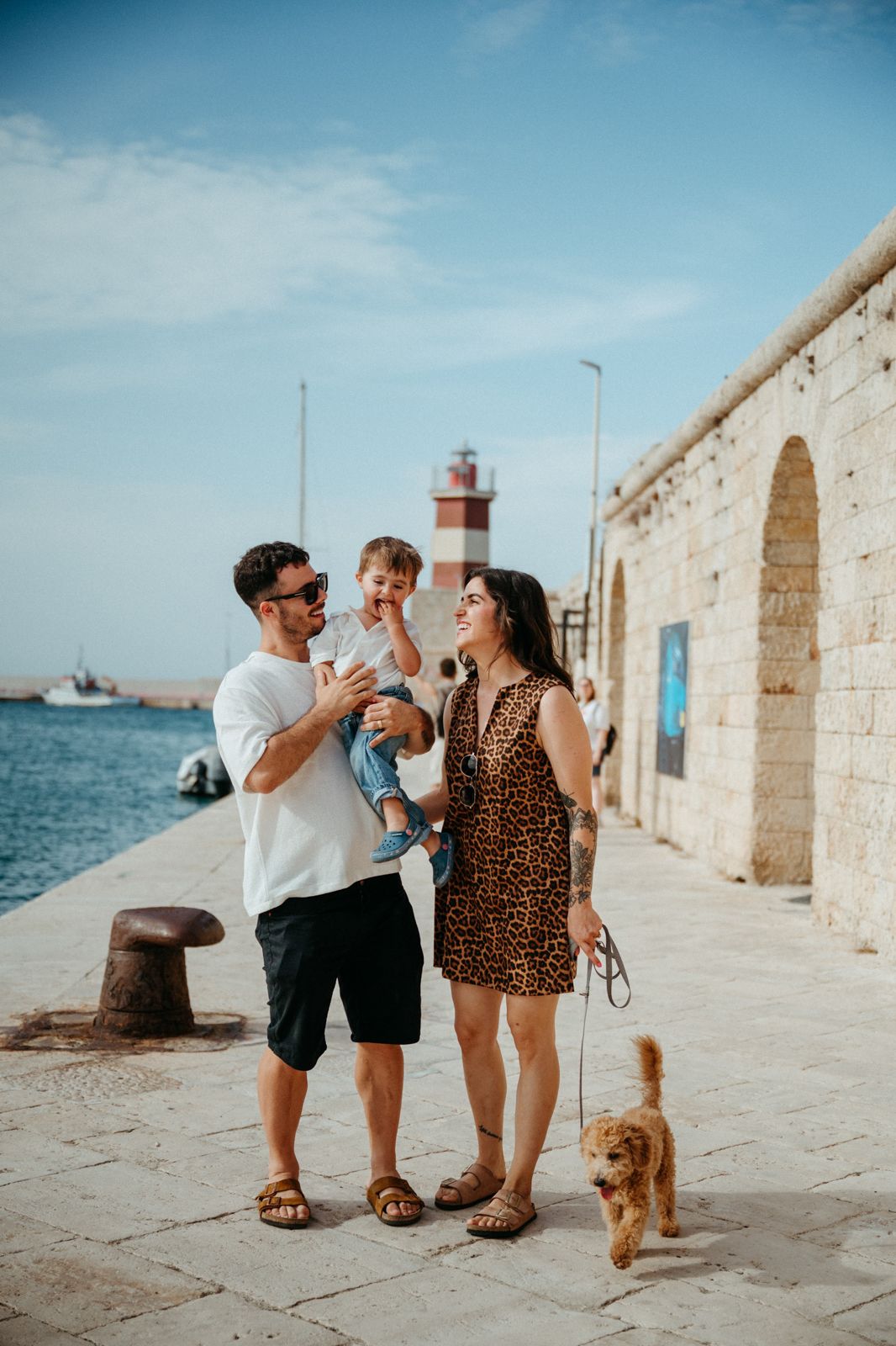 Family with toddler and golden dog strolling along sunny Mediterranean harbor with red lighthouse.