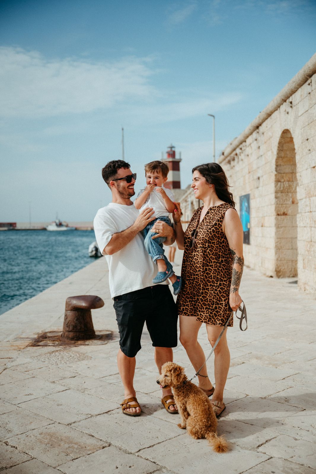 Family with toddler and golden doodle dog walking along sunny Mediterranean harbor promenade with lighthouse.