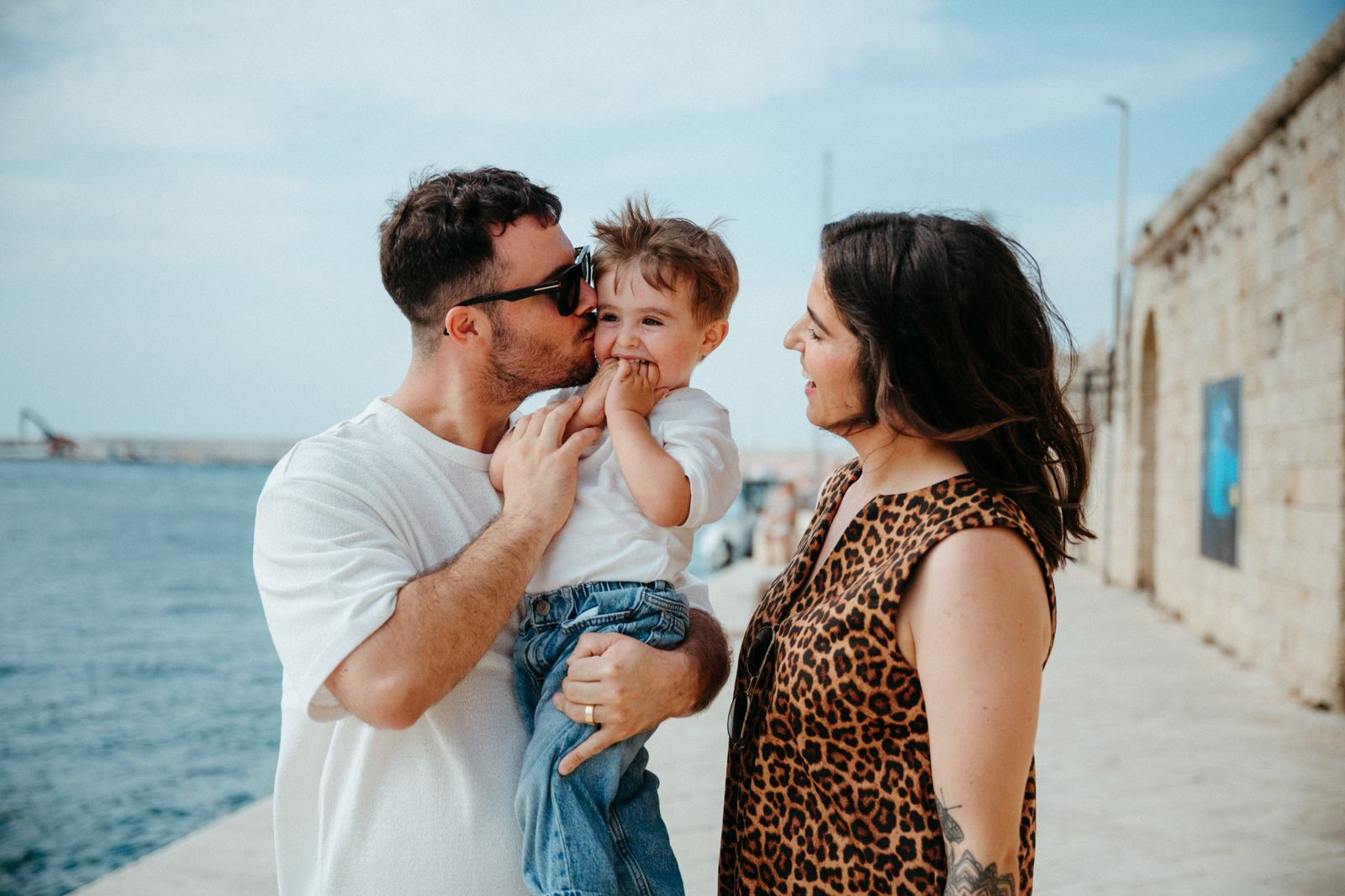 Family of three enjoying a sunny seaside day, parents smiling at toddler by the ocean.