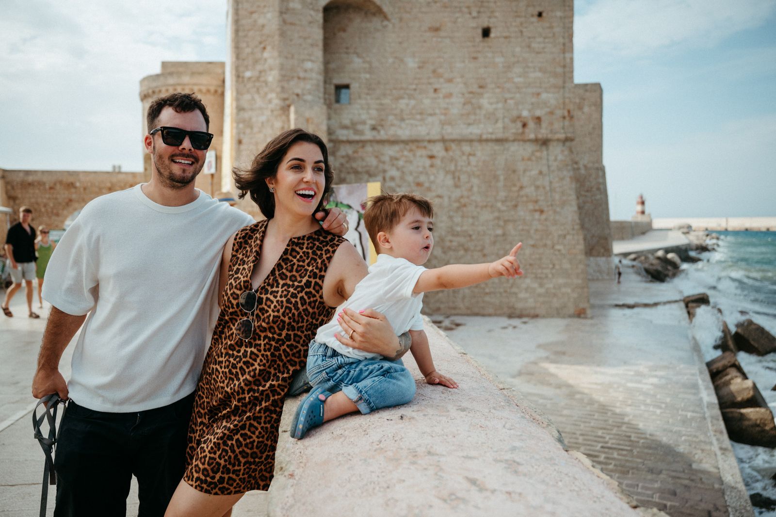 Happy family with baby posing by ancient stone fortress near Mediterranean seaside on sunny day.