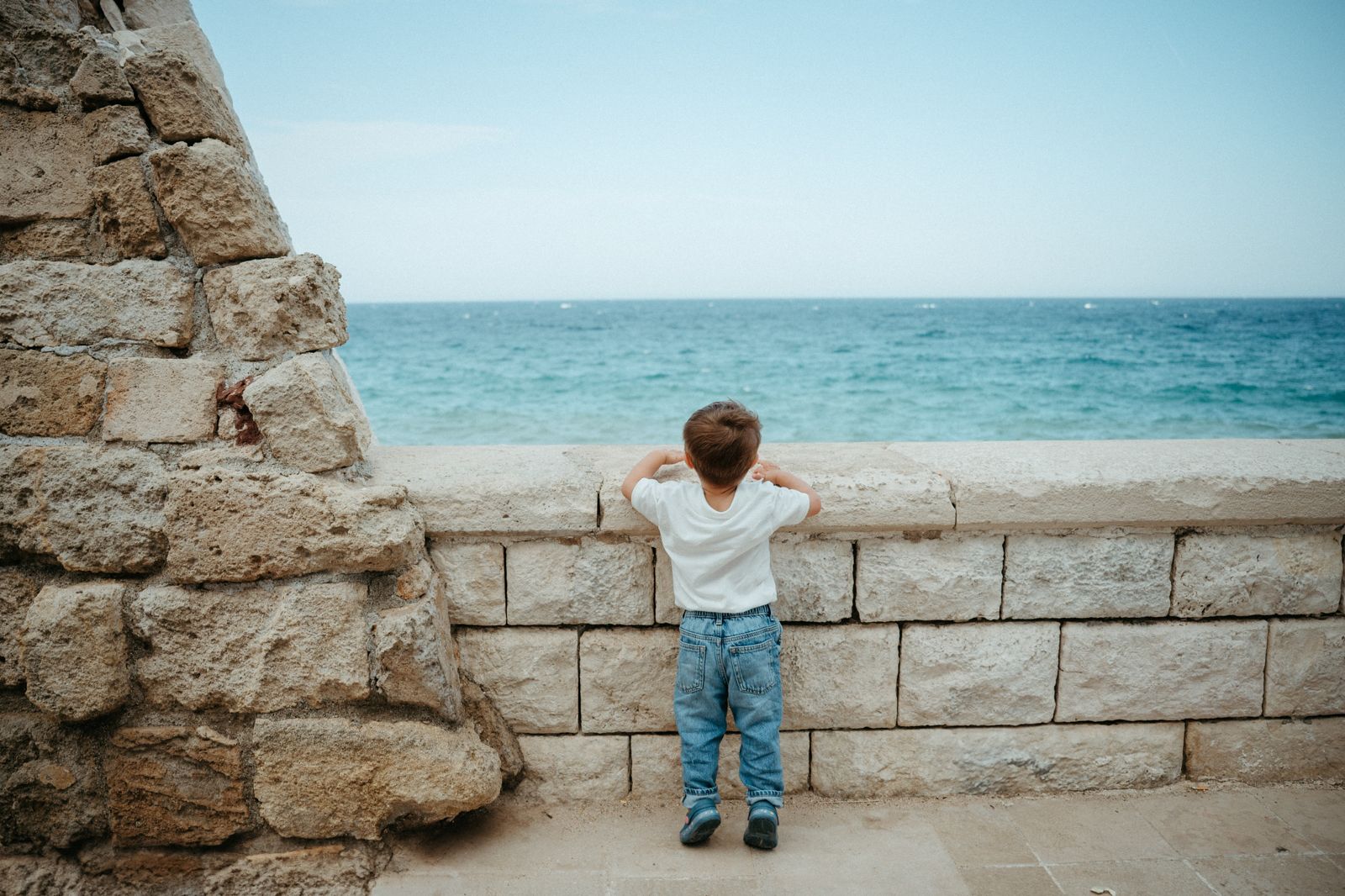 Young boy standing on tiptoes looking over a stone wall at the blue Mediterranean sea.