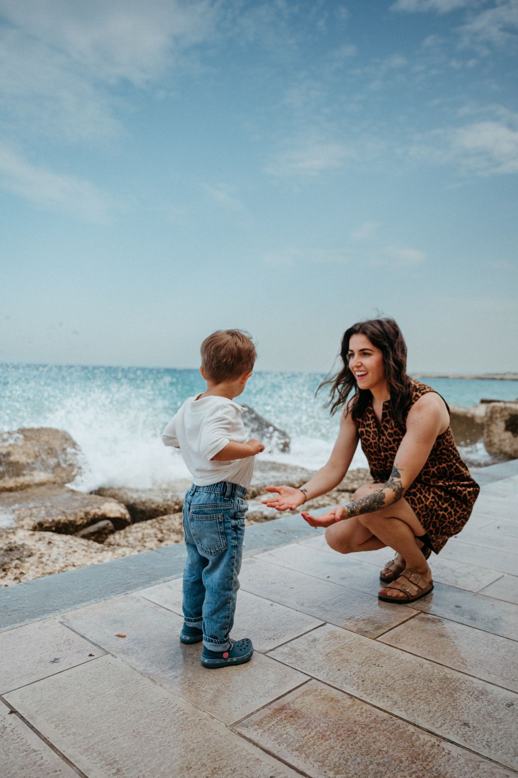 A smiling mother crouches down to greet her toddler son by the ocean on a sunny day.