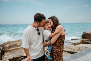 Family of three sharing a joyful moment by the ocean, parents kissing baby on rocky Mediterranean coast.