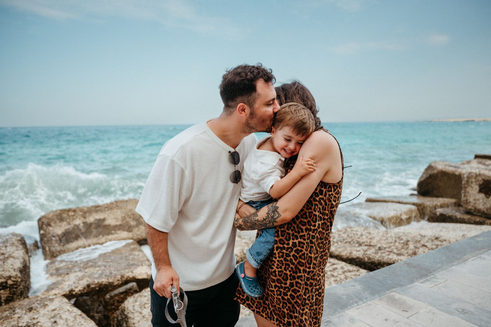 Couple kissing while holding their young child near rocky Mediterranean coastline.