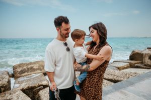 Happy couple holding toddler on rocky seaside with turquoise ocean waves in background.