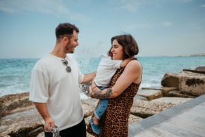 Happy young family with baby enjoying a sunny day by the turquoise sea on a rocky coastline.