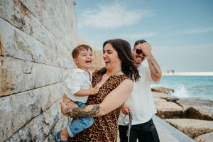 Family laughing together by seaside stone wall, mother in leopard dress holding toddler, father celebrating behind them.