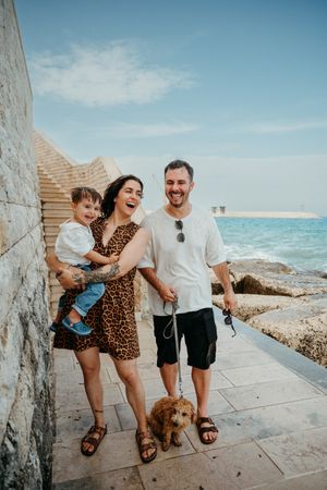 Happy young family with baby and golden doodle dog laughing by seaside stone wall on sunny day.
