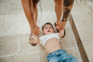 Two adults hold a laughing baby lying on a stone floor, with a tattoo visible on one arm.