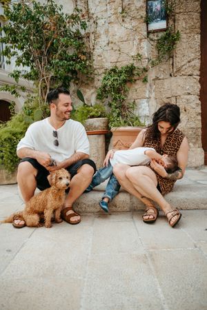 Family with golden puppy relaxing on stone steps outside rustic Mediterranean building.
