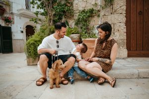Young family sitting on cobblestone street with golden dog, parents smiling at baby in sunny European town.