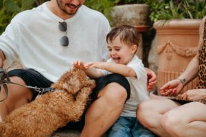 Happy toddler laughing while playing with a fluffy golden doodle dog outdoors.