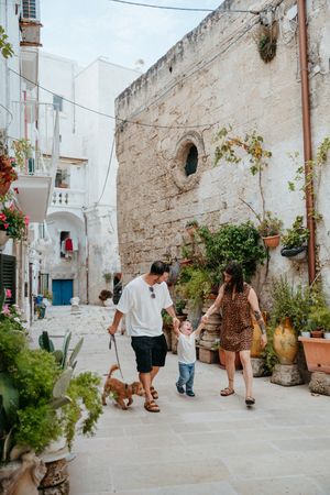 Family with toddler and dog strolling through charming white-washed Italian alleyway with potted plants.