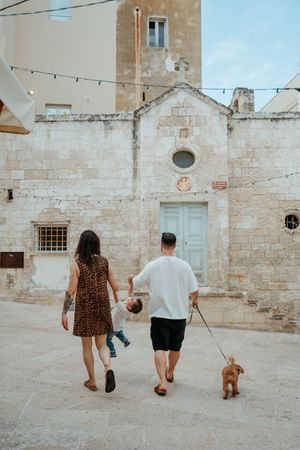 A family with a toddler and a dog walks toward an old stone church in a European town.