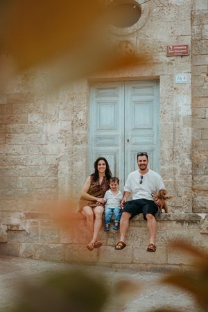 Family of three sitting on stone steps with a dog in front of a rustic blue door in a Mediterranean town.