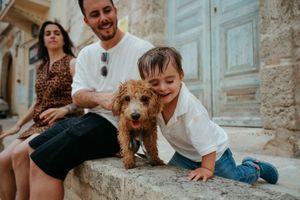 A happy family with a young boy and man petting a fluffy golden doodle puppy on stone steps in a European town.