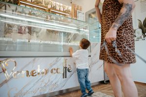 Young toddler boy looking at display case inside Veneto Cafe with tattooed mother nearby.