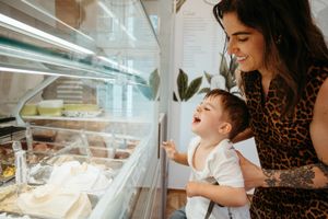 Young mother in leopard print dress holding toddler pointing at ice cream display case in a shop.
