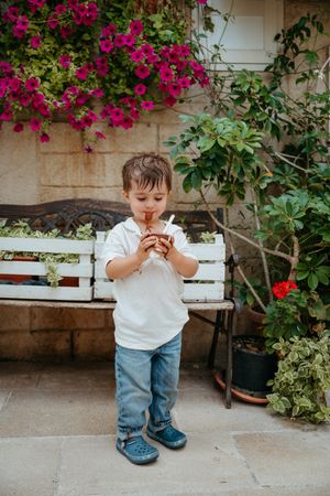 Cute little boy in white shirt and jeans drinking from a cup outdoors near colorful flowers.