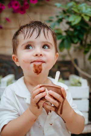 Messy toddler boy with chocolate ice cream on his face holding a cup with a straw outdoors.