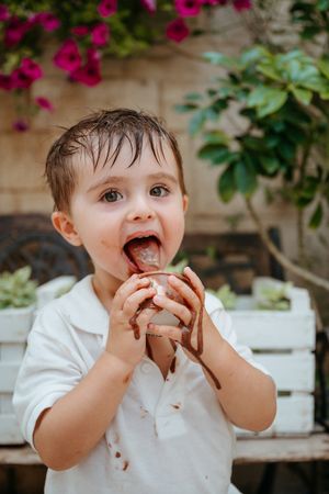Toddler boy in white shirt eating chocolate ice cream with messy hands outdoors near pink flowers.