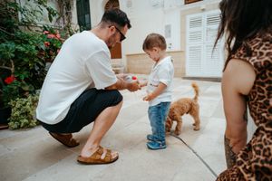 Father crouching to give toy to toddler son outdoors while fluffy dog plays nearby.