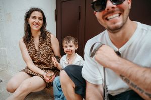 Smiling family with young child sitting outdoors, man with tattoo and sunglasses in foreground.