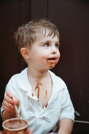 Young boy with chocolate smeared on his face and shirt holding a spoon and cup indoors.