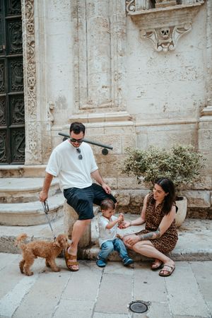 Family with toddler and fluffy dog relaxing on stone steps outside ornate historic building on sunny day.