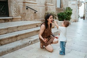 A woman in a leopard print dress kneeling to interact with a happy toddler on a cobblestone street.