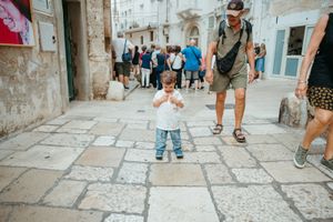 A toddler stands on a cobblestone street in a European town while an adult man walks nearby.