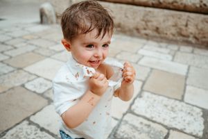 Adorable toddler boy with chocolate-stained white shirt eating a treat on a stone patio.