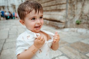 A messy toddler boy in a white shirt smiles while eating ice cream outdoors in a historic stone plaza.