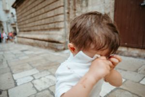 Young toddler boy in white shirt covering face with hands on cobblestone street in old European town.