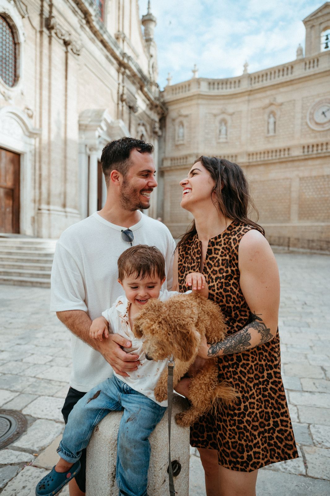 Happy family with young son and fluffy dog smiling together outside a historic European cathedral.