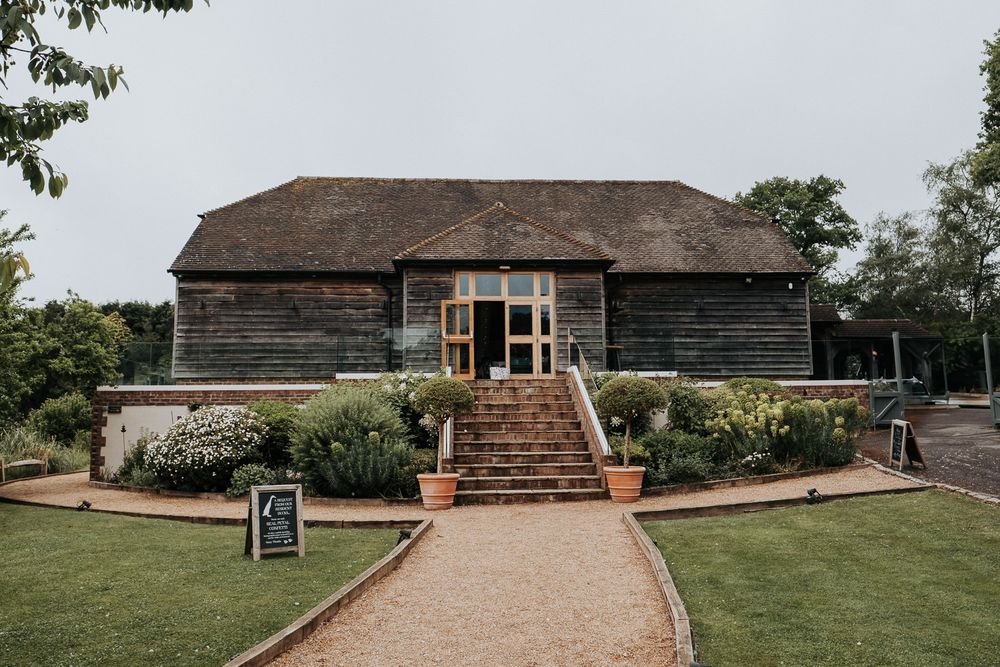 Rustic wooden barn venue with brick steps, potted plants, and a gravel pathway leading to the entrance.
