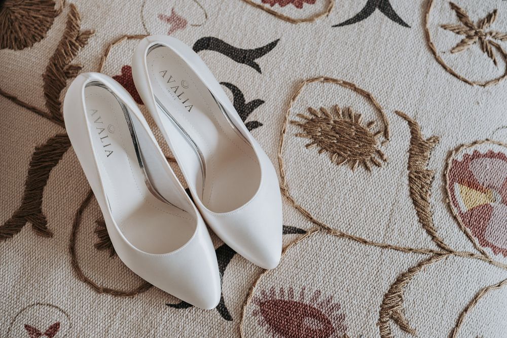 White satin pointed-toe bridal heels by Valia placed on a floral patterned carpet.