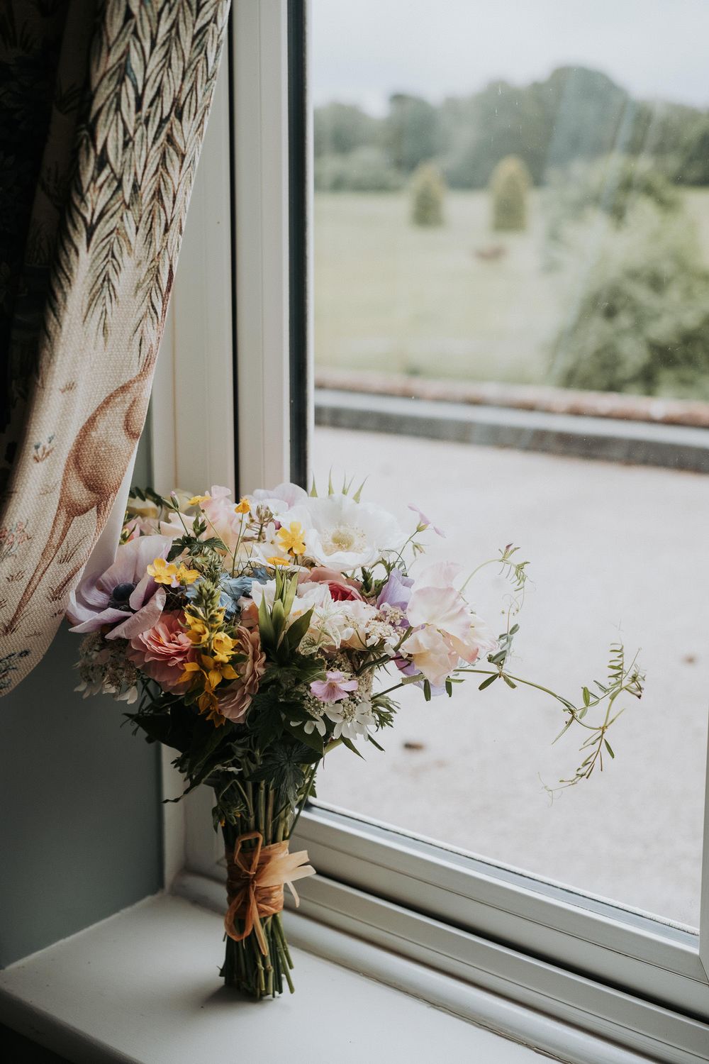 Colorful wildflower wedding bouquet tied with twine sitting on a windowsill overlooking a green countryside.