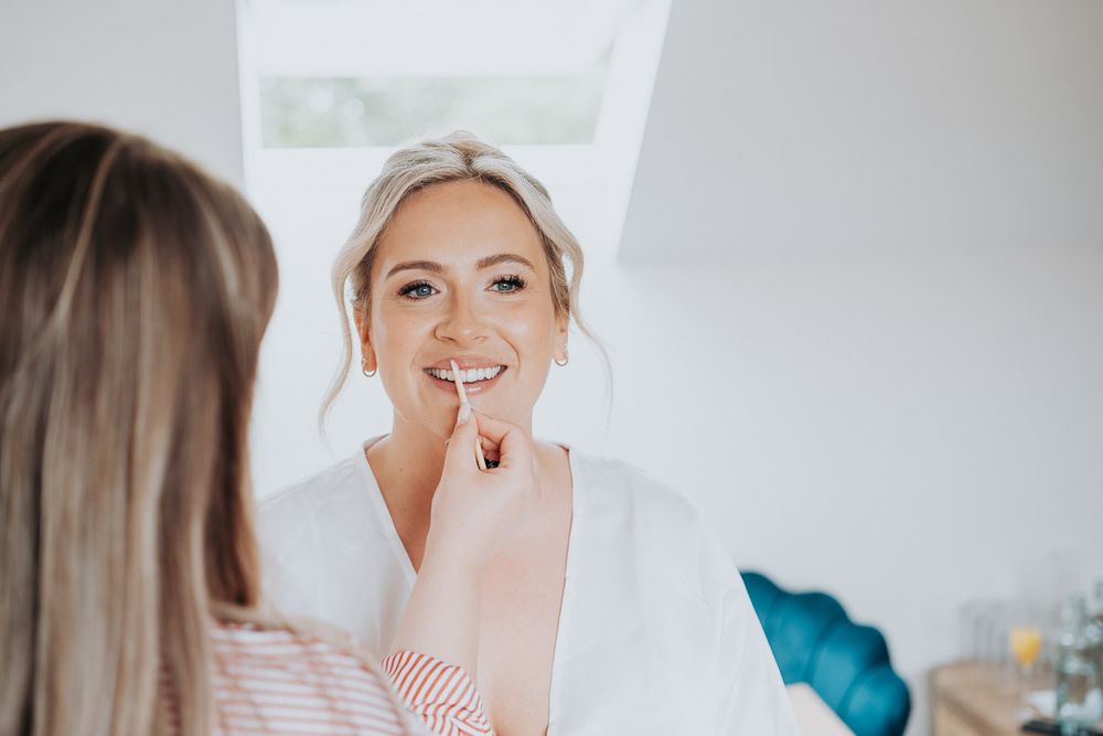 Makeup artist applying lipstick to a smiling blonde bride in a white robe during wedding preparations.